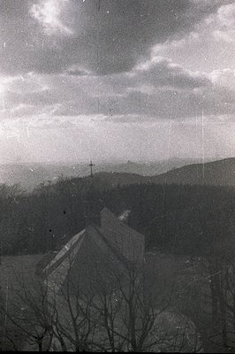 Striking black and white photograph depicting a church atop a hill overlooking a forested valley. A prominent cross crowns th...