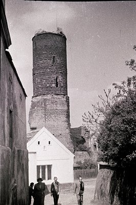 Tall, weathered brick tower rises above a whitewashed building. Three men in suits walk along a path. Appears to be a courtya...