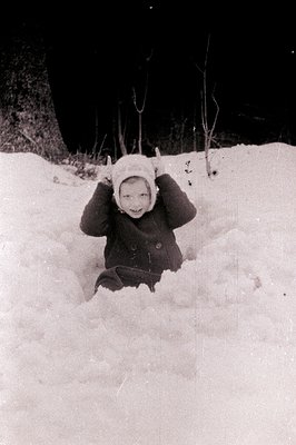 A child bundled in a hooded coat and mittens playfully poses, partially submerged in a snowdrift. The wintry scene features m...