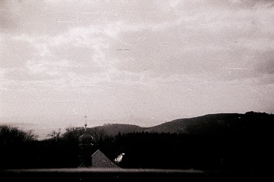 An Orthodox church with prominent onion domes sits on a gentle slope, silhouetted against a dark treeline and cloudy sky. App...