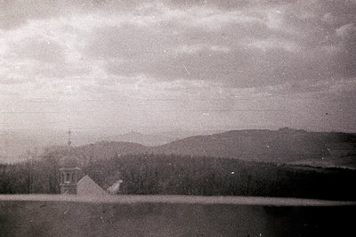 Ornate, domed Orthodox church sits on a gentle rise, overlooking a dark, still body of water. Dense forest flanks the water's...