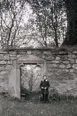 Ruined stone structure frames a dense landscape, with a person in a dark coat and hat standing prominently. The weathered sto...