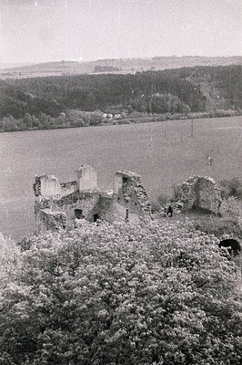 Overlooking a river or lake, remains of a stone structure stand amidst dense foliage. Architectural ruins suggest a medieval ...