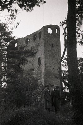 Overgrown ruins of a stone tower, likely a former defensive structure. Two men in suits and hats stand near the base, providi...