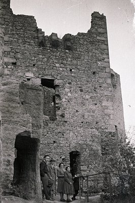 A black and white photograph depicts four people posing in front of a ruined stone tower, likely a fortification. Three indiv...