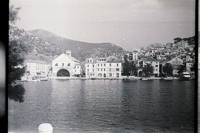 Coastal townscape featuring waterfront buildings and a partially visible archway. Buildings exhibit traditional Mediterranean...