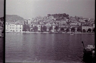 Coastal townscape with tiered buildings and palm trees ascending a hillside overlooking the sea. Boats are docked along the w...