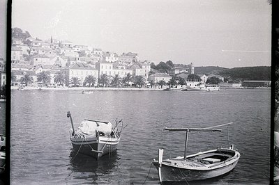 A calm coastal scene features two traditional fishing boats moored in the foreground, with a hillside town rising steeply in ...