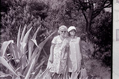 Two women in vintage patterned dresses and headwear stand outdoors, framed by tropical foliage. Likely a resort or vacation s...
