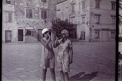 Two women stand smiling in a cobbled square, peering through binoculars. One wears a checked dress-suit; the other a checked ...