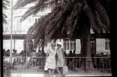 Two women in summer dresses stand before a large palm tree and a café with outdoor seating. The image, likely from the 1960s...