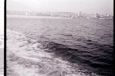 Black and white image captures a ship's wake churning the water, with a coastal cityscape in the distance. Appears to be a po...