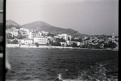 A coastal town clings to a hillside, with a stone wall defining the waterfront. Buildings exhibit varied architectural styles...