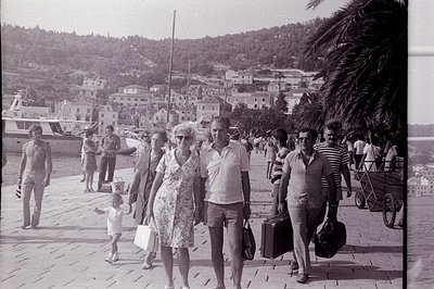 A group strolls along a promenade, likely a seaside resort, with boats anchored in the harbor. Terraced hillside housing and ...