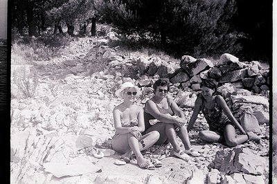 Three young women, seated on a rocky outcrop, wearing swimwear. A low stone wall forms a backdrop. Likely a resort or coastal...