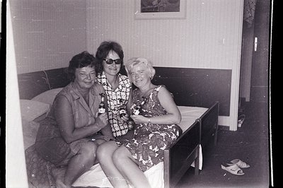 Three women seated on a bed in what appears to be a hotel room, each holding a bottle of beer. Floral patterned dresses and 1...