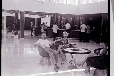 A leisure scene within a 1970s-era hotel lobby. A man and woman, dressed stylishly, relax in rattan chairs beside a circular ...
