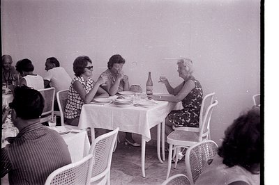 Three women seated at a table covered with a white linen cloth, enjoying drinks and conversation. The setting appears to be a...
