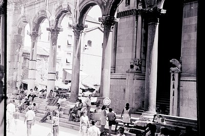 A black and white photo shows a grand, classical arcade with numerous tall columns and statues. People are seated and standin...