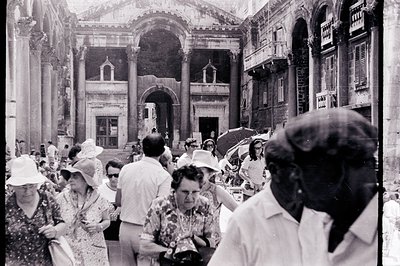 A crowd of tourists traverses a stone courtyard flanked by classical architecture, possibly Roman ruins. People wear 1960s-70...