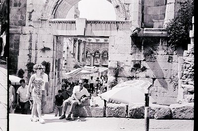 A black & white architectural view showcasing a bustling public space framed by aged stone ruins. A woman in a patterned dres...