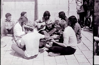 Group of young men, likely musicians, gathered outdoors. A guitarist sits center, while others hold instruments or listen. Si...
