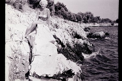 A woman in a white bandeau swimsuit poses on a rocky cliff overlooking the ocean. The shoreline features rugged, light-colore...