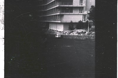 A 1960s or 70s view of a modernist apartment building. A small, light-colored car sits in a sloped, landscaped area below the...
