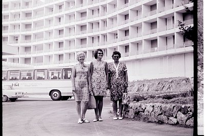 Three women in patterned dresses and sunglasses stand outdoors in front of a large, modernist hotel. A bus labeled "Hotel Med...
