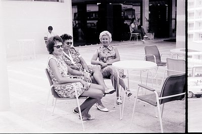 Two women sit in patio chairs facing the camera, both in patterned, short-sleeved dresses and sunglasses, a style typical of ...