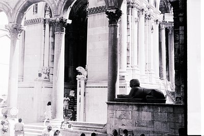 Dramatic black and white image showcasing grand, classical architecture: towering marble columns, a stone lion statue, and ti...