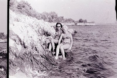 Two women in swimwear pose on a rocky outcrop overlooking a seaside landscape. One wears a patterned dress, the other a flora...