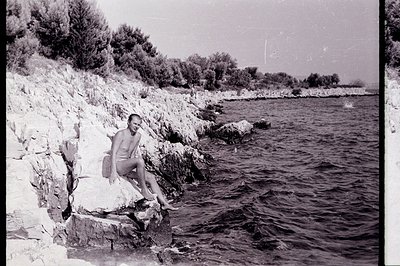 A bare-chested man sits on a rocky ledge overlooking a dark seascape. Jagged limestone cliffs and sparse Mediterranean vegeta...
