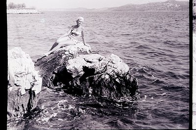 A young woman in a vintage swimsuit poses on a rocky outcrop overlooking a tranquil seaside. Possibly 1960s or 70s, the compo...