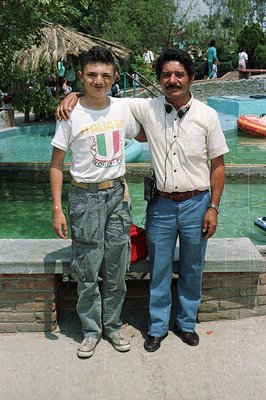 A young man and older gentleman, posed shoulder-to-shoulder, stand beside a recreational pool with visible water slides and i...