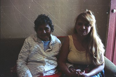 Two individuals sit side-by-side on a cushioned bench, a red wall partially visible behind them. The person on the left wears...