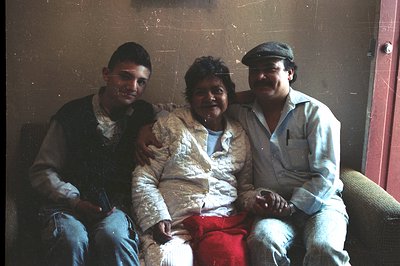 Three individuals sit closely together—a young man, a woman, and an older man—in an interior setting. The men flank the woman...