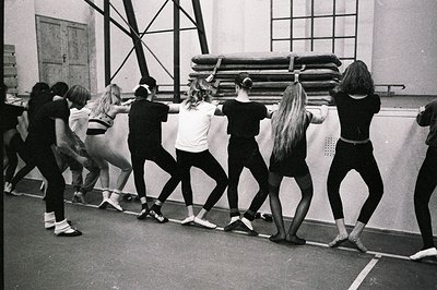 A line of young ballet students, facing away, practice near a barre. Stacked gymnastics mats lean against the wall of a gymna...