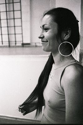 A young woman in a simple tank top and large hoop earring is captured in profile. She appears to be gazing out of a window, h...
