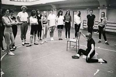 A dance instructor demonstrates a movement, seated on a chair, to a group of teenage ballet students in a rehearsal space. Th...