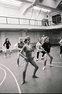 A group of young women in a dance studio during a movement or aerobics class. Several participants wear leotards; others in c...
