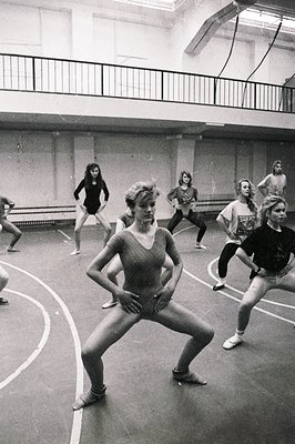 Black and white image capturing a dance class in a gymnasium. A young woman in a leotard demonstrates a pose while several ot...