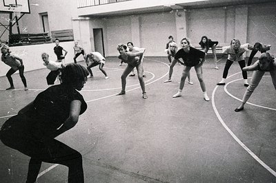 Group of young women in athletic wear participating in a dance or fitness class. Instructor leads warm-up exercises in a gymn...