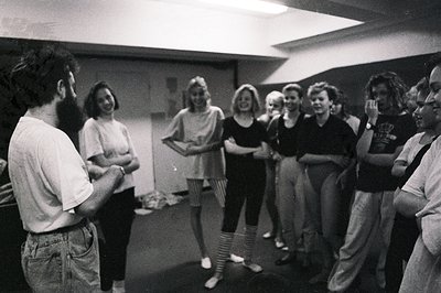 A group of young adults gathers in a brightly lit, sparse room, possibly a rehearsal or workshop space. A man with a beard is...