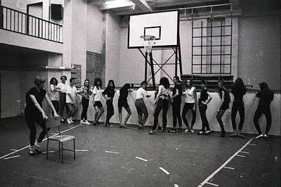 A group of young women in leotards and tights practice ballet in a gymnasium. A female instructor gestures towards the studen...