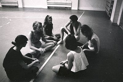 Six young women sit in a circle on a polished floor, possibly a dance studio or gymnasium. The scene features plain walls, a ...