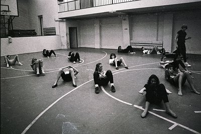 A group of young people, mostly women, are casually seated and reclining on a marked indoor athletic court. The scene evokes ...