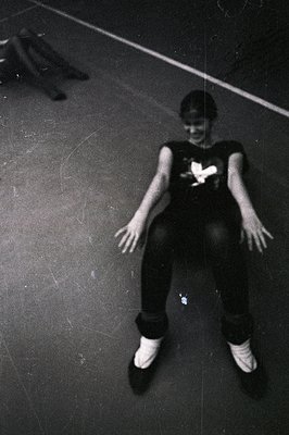 A young person sits cross-legged on a dark floor, wearing a graphic tee and sneakers. A blurred figure stands in the backgrou...