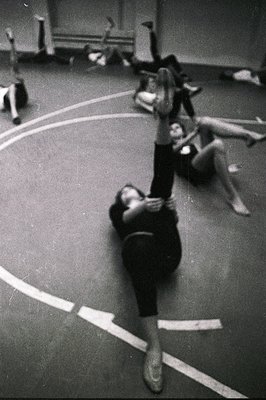 A ballet studio scene captures a dancer in mid-motion, likely rehearsing a lift, with other dancers resting or preparing in t...