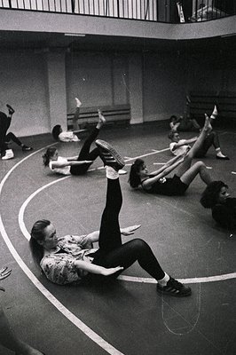 A black and white image shows a modern dance class in progress. Several students are performing exercises, one prominently li...